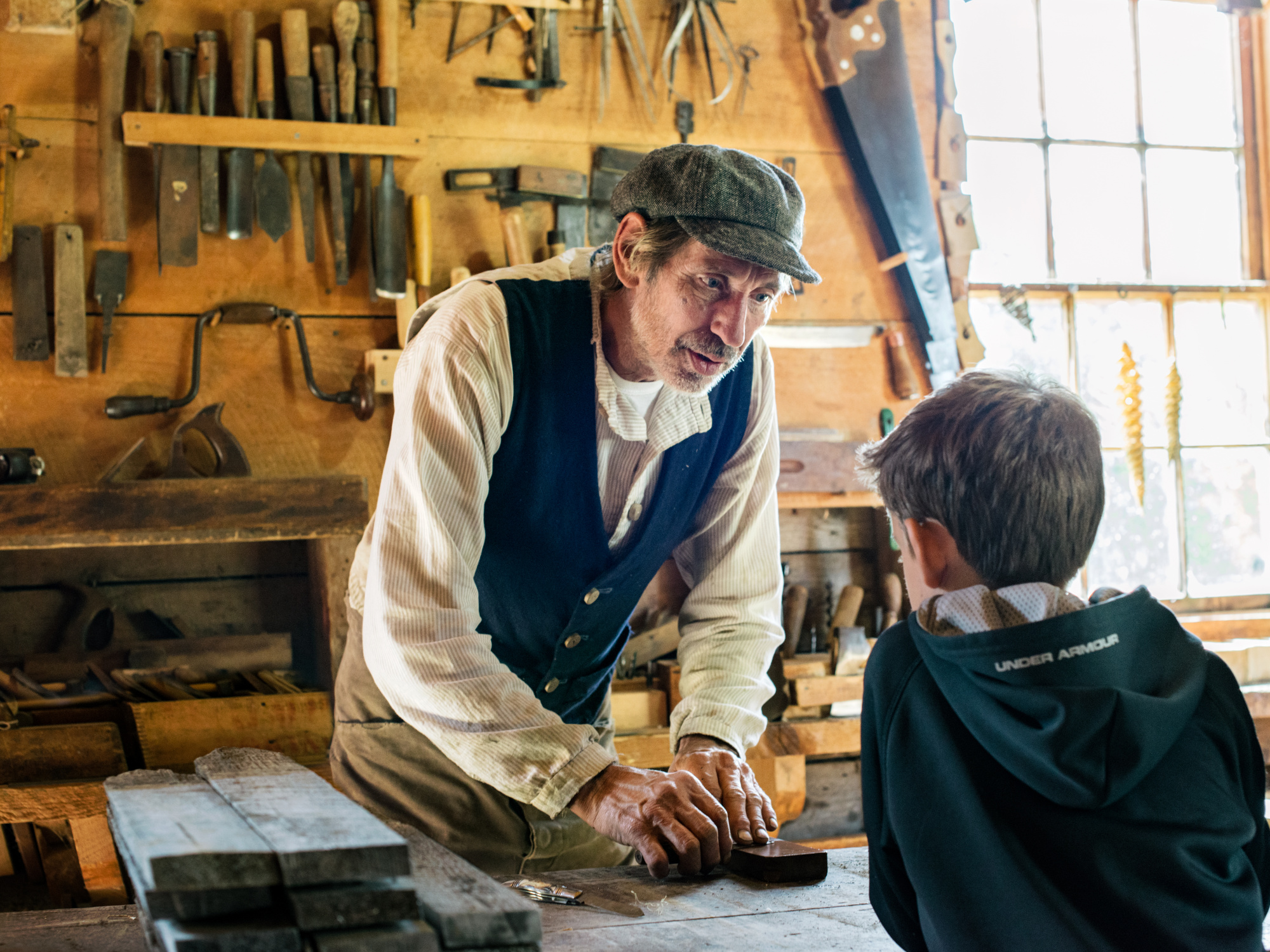 Costumed interpreter, a man behind the counter of the Farm Workshop speaking to a young boy. 