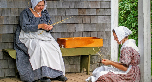 Two costumed interpreters on the porch of Rosebank Cottage making straw hats.