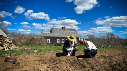 Costumed interpreters, a woman and a man planting seeds in a field at Ross Farm.