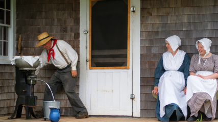 Two female costumed interpreters sit on the porch of Rosebank cottage watching a male costumed interpreter churn butter.
