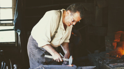 A blacksmith in historical attire works inside a dimly lit workshop. The man wears a light-colored, loose-fitting collared shirt with suspenders, dark trousers, and eyeglasses. Leaning forward, he holds a hammer down against a piece of glowing hot metal resting on a large metal anvil. To the right, a brick forge glows with a bright orange fire, while a multi-pane window on the left lets in natural daylight, illuminating the workspace.