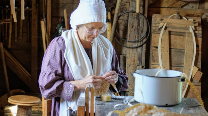 A woman dressed in historical 19th-century attire sits inside a wooden workshop, focused on a handcrafting task. She wears a purple patterned long-sleeved dress, a white apron, a white shoulder wrap, a ruffled white bonnet, and wire-rimmed eyeglasses. She holds and manipulates a woven, textured material in her hands. In the foreground, a white enamel pot with a black rim sits on a workbench alongside woven shavings and craft supplies. The rustic background features wooden walls lined with tools.