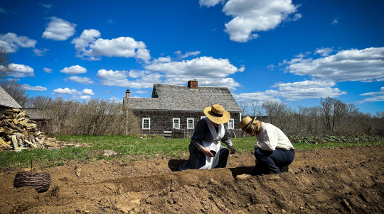 Costumed interpreters, a woman and a man planting seeds in a field at Ross Farm.