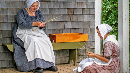 Two costumed interpreters on the porch of Rosebank Cottage making straw hats.
