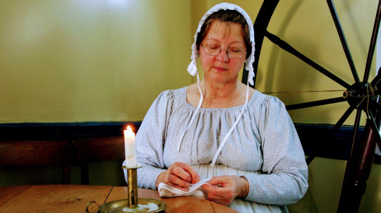 Costumed interpreter, a woman sewing at a table.