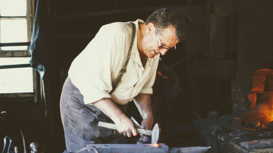 A blacksmith in historical attire works inside a dimly lit workshop. The man wears a light-colored, loose-fitting collared shirt with suspenders, dark trousers, and eyeglasses. Leaning forward, he holds a hammer down against a piece of glowing hot metal resting on a large metal anvil. To the right, a brick forge glows with a bright orange fire, while a multi-pane window on the left lets in natural daylight, illuminating the workspace.