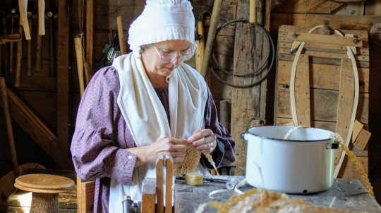 A woman dressed in historical 19th-century attire sits inside a wooden workshop, focused on a handcrafting task. She wears a purple patterned long-sleeved dress, a white apron, a white shoulder wrap, a ruffled white bonnet, and wire-rimmed eyeglasses. She holds and manipulates a woven, textured material in her hands. In the foreground, a white enamel pot with a black rim sits on a workbench alongside woven shavings and craft supplies. The rustic background features wooden walls lined with tools.